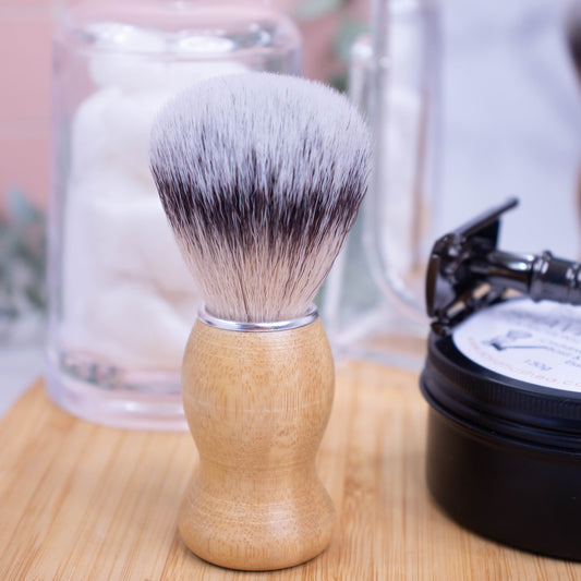 A shaving brush stands upright on a wooden surface next to black tin of shaving soap with a razor.   The Shaving brush has a wooden handle with a silver collar and bristles that are layered in shades of cream, black then grey.
