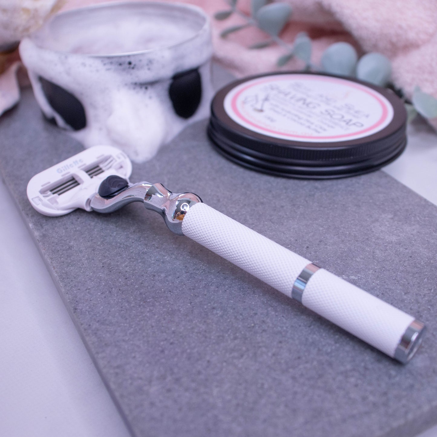 A Razor with textured white handle and silver trim with Gillette cartidge lies on a stone tray next to an open tin of shaving soap with a dense foam flowing over the edges.