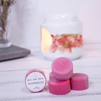 Four cylindrical shaped Melt Pucks in pink/red are stacked up on each other in pyramid form with the soy and shea logomark stamped on the tops.  To the left is a fourth melt puck that rests on its side with a label identifying it as Watermelon.  These sit on a wooden surface with a  lit fragrance warmer in the backgroung along with a vase of flowers.