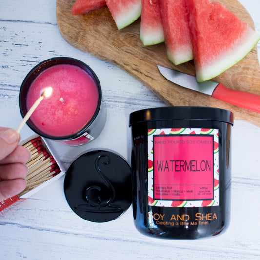 A flat lay photo showing a large black jar lying on its side on a white wooden surface below a wooden board with slices of watermelon.  To the left of the jar is a smaller candle standing upright and filled with bright red wax.  A hand reaches in with a lit match.