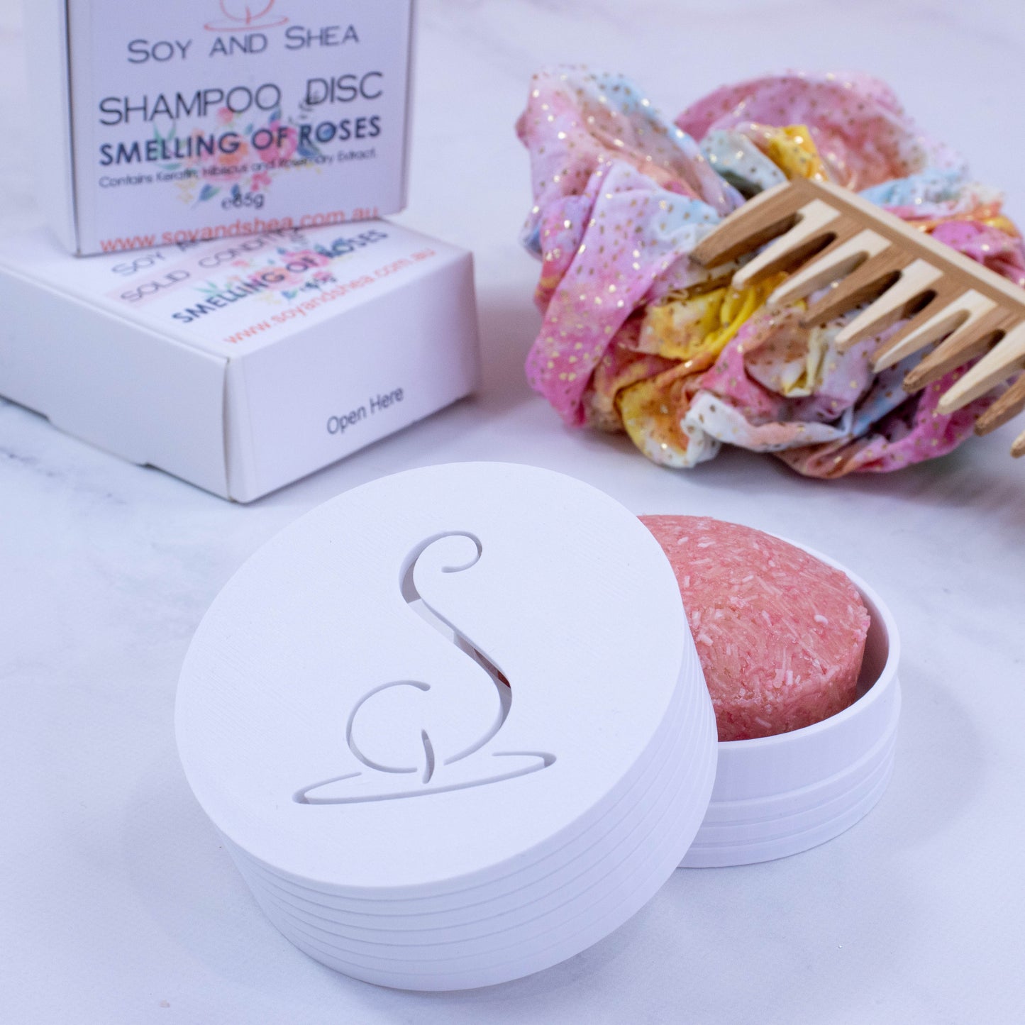 A white cylindrical pot with the Soy and Shea logomark cut out in the top, sits on a white surface.  The lid has been removed and is resting on the side of the base which holds a solid shampoo bar.  In the background stands the shampoo packaging next to a wooden comb and colourful scrunchie with shiny gold dots.