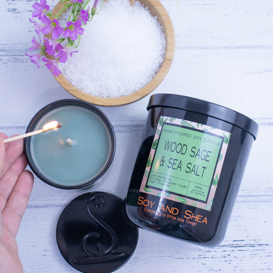 A flat lay photo showing a large black jar lying on its side on a white wooden surface next to a wooden bowl filled with salt and a sprig of purple flowers.  To the bottom of the jar is a smaller candle standing upright and filled with dark green wax.  A hand reaches in with a lit match.