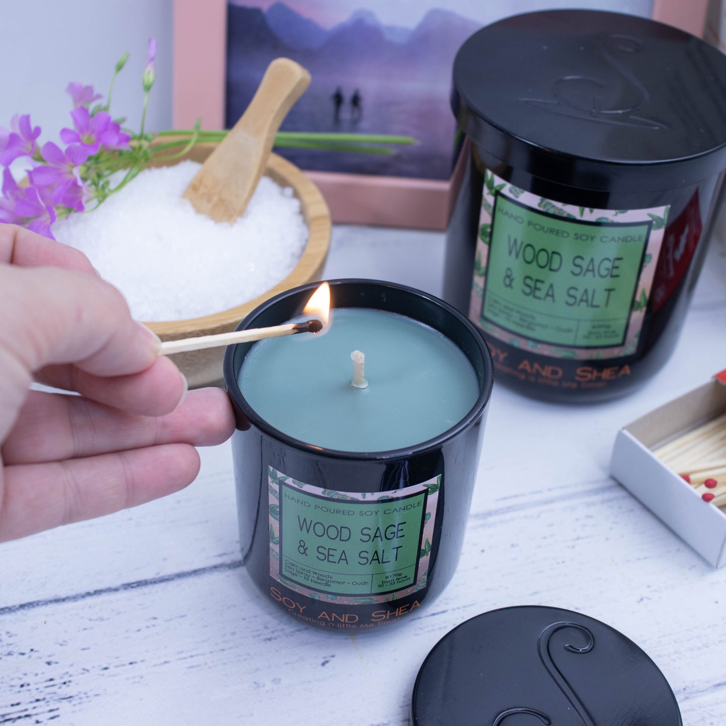 A hand reaches in with a lit match to light a green candle in a black glass jar with product label.  Behind is a larger candle with a black metal lid with logomark embossed into it.   This sits next to a wooden bowl filled with salt and a sprig of purple flowers and a photoframe with watercolour painting.