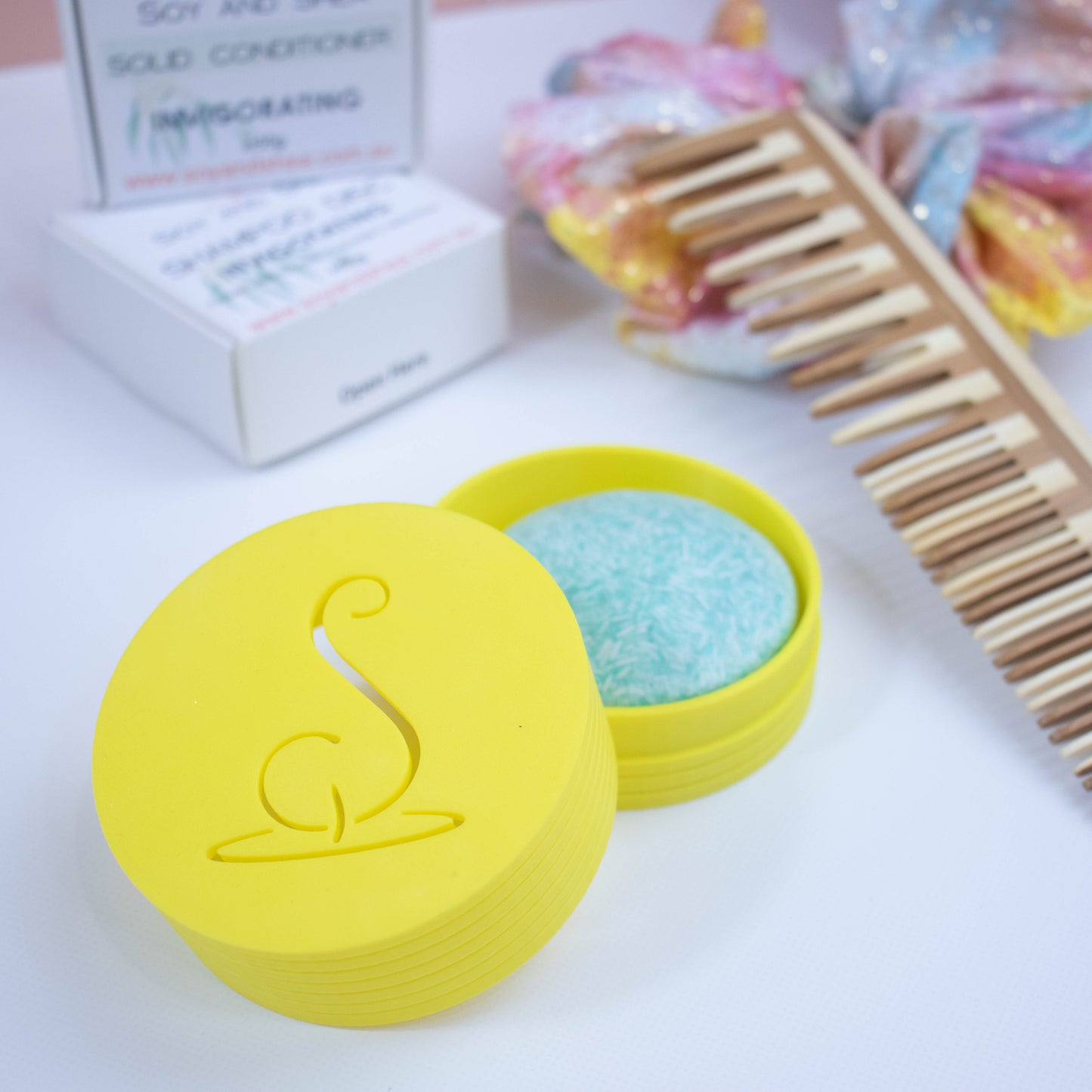 A bright yellow cylindrical pot with the Soy and Shea logomark cut out in the top, sits on a white surface.  The lid has been removed and is resting on the side of the base which holds a solid shampoo bar.  In the background stands the shampoo packaging next to a wooden comb and colourful scrunchie with shiny gold dots.